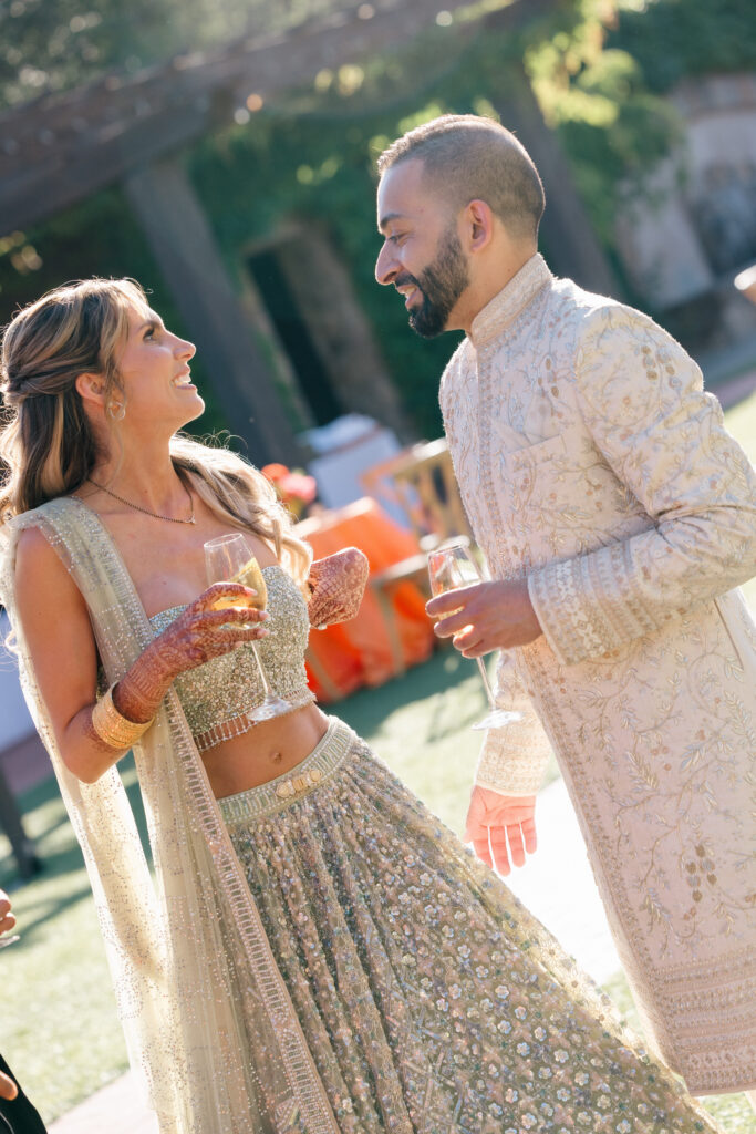 A bride and groom laugh together over glasses of champagne during their Sangeet celebration at Estate Yountville in Napa Valley, the bride in a gold embroidered lehenga with henna covered hands, photographed by Suzanne Karp Photography