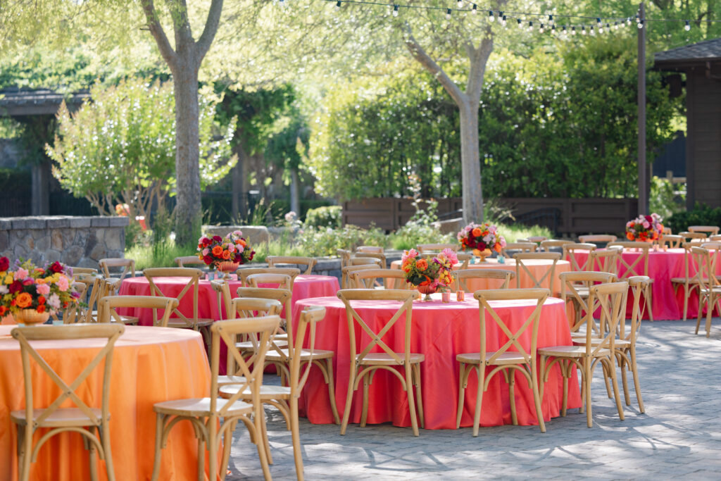 A vibrant outdoor Sangeet celebration at Estate Yountville with coral and orange linen tables, crossback chairs, and jewel-toned floral centerpieces set beneath mature trees in Yountville, Napa Valley, photographed by Suzanne Karp Photography