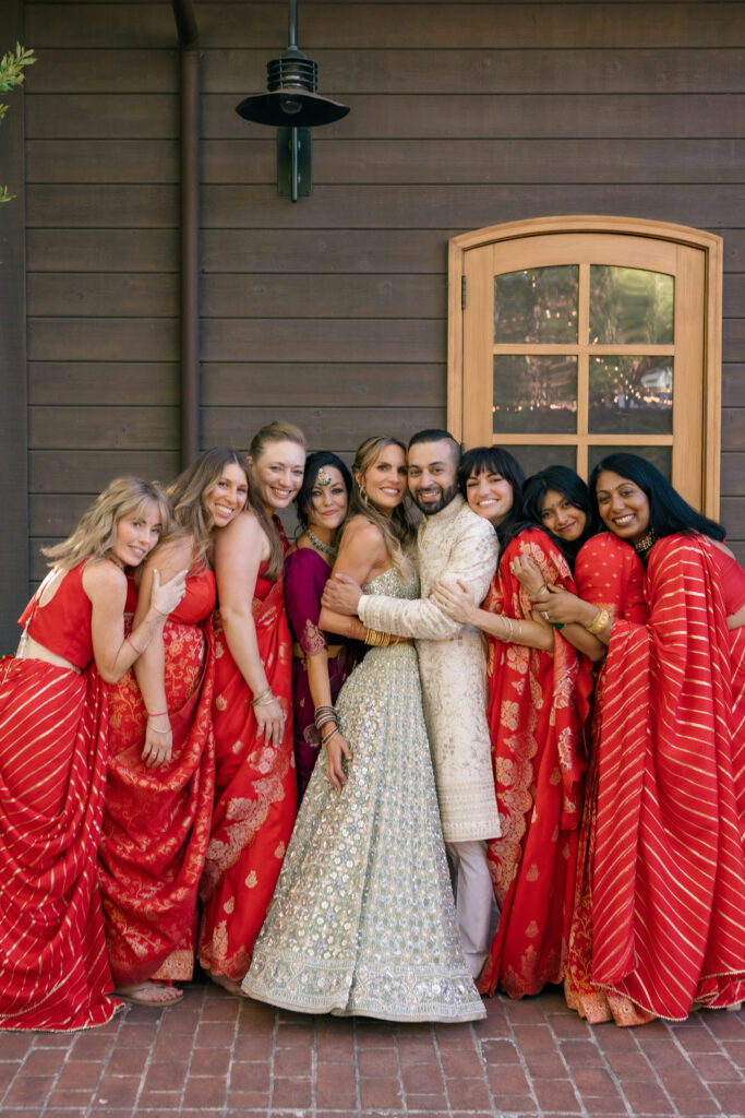 A bride in a gold embroidered lehenga poses with her bridal party dressed in vibrant red banarasi sarees outside Estate Yountville during a Sangeet celebration in Napa Valley, photographed by Napa Valley wedding photographer Suzanne Karp