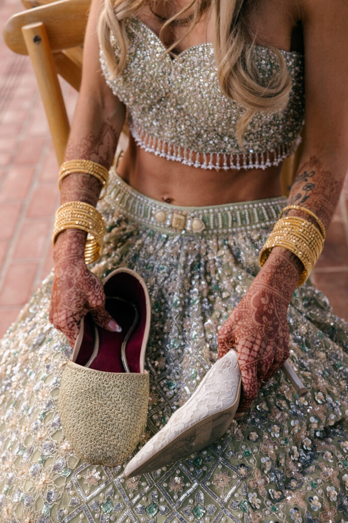 A close up detail of a bride's henna covered hands and gold bangles holding embellished bridal shoes during the Sangeet celebration at Estate Yountville in Napa Valley, photographed by Suzanne Karp Photography