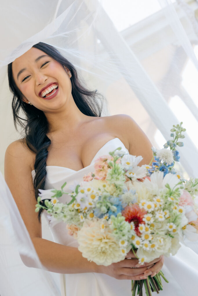 A bride laughs joyfully while holding a wildflower bouquet of daisies, blue delphinium, and peach blooms in soft window light during her wedding day preparations, photographed by Napa Valley wedding photographer Suzanne Karp