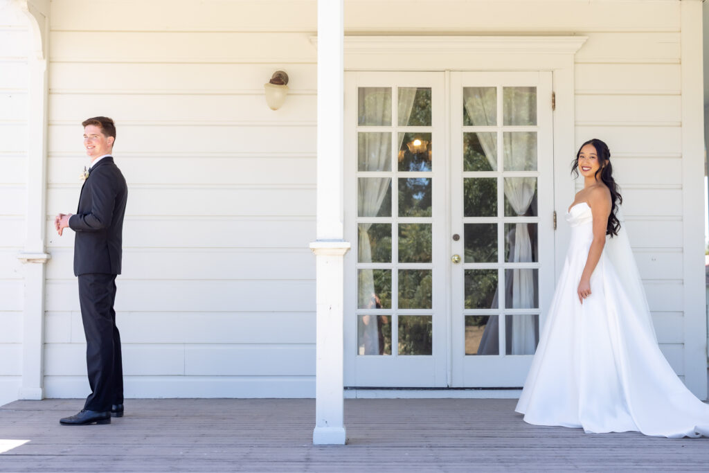 A groom stands with his back turned waiting for his first look while his bride in a strapless ballgown smiles on the porch of a white Victorian farmhouse during a Northern California wedding, photographed by Suzanne Karp Photography