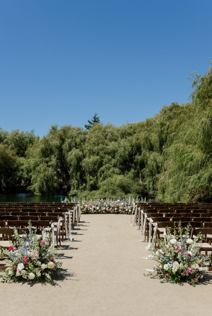 A wide view of an outdoor wedding ceremony setup with wooden chairs lining a sandy aisle, colorful floral arrangements, and weeping willow trees beside a peaceful waterway under a deep blue sky in Northern California, photographed by Suzanne Karp Photography