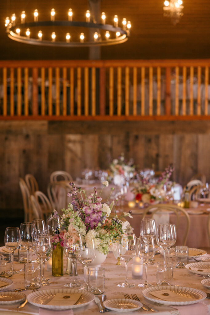 An intimate barn wedding reception tablescape with blush linen tables, mixed wildflower centerpieces, wine bottle vases, and glowing candlelight beneath a circular chandelier in a Northern California barn venue, photographed by Suzanne Karp Photography