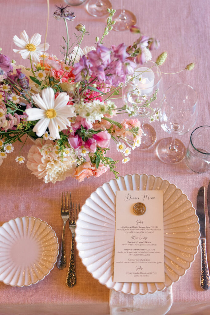 A close up wedding place setting detail with a scalloped white plate, gold wax seal menu card, and a vibrant wildflower centerpiece of daisies, sweet peas, and cosmos on a blush linen table, photographed by Napa Valley wedding photographer Suzanne Karp