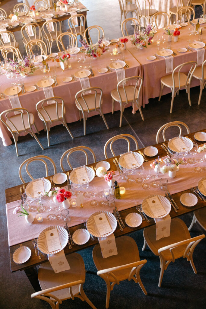 An aerial view of long blush linen reception tables set with gold bistro chairs inside a warmly lit barn wedding venue in Northern California, photographed by Suzanne Karp Photography