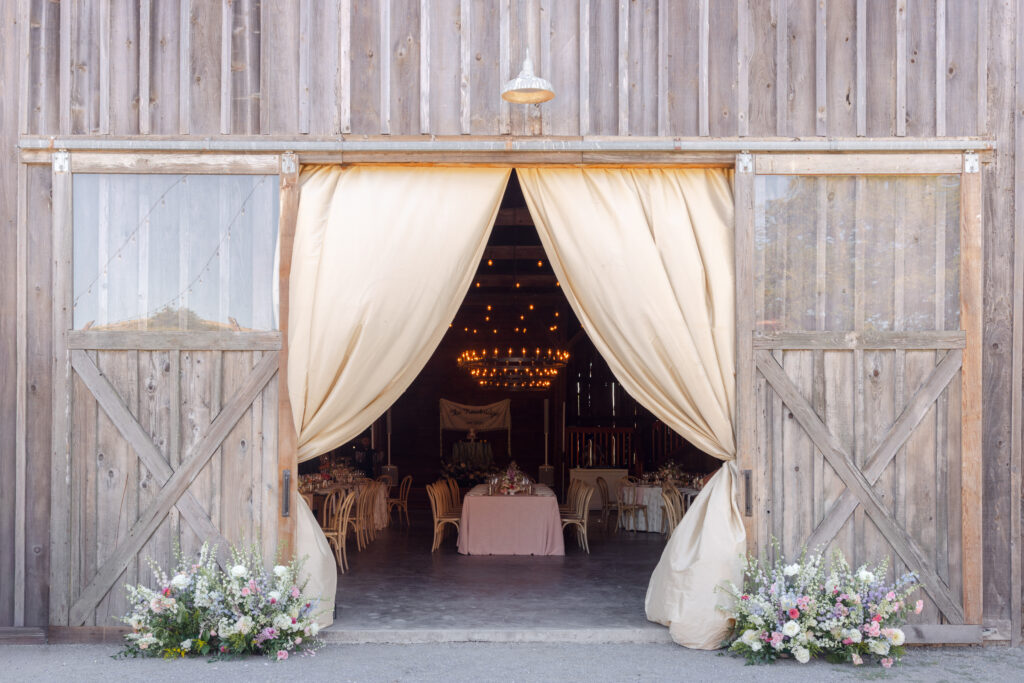 A rustic barn wedding reception venue with sliding wood doors draped in cream fabric, wildflower arrangements flanking the entrance, and a warmly lit chandelier interior visible beyond, photographed by Napa Valley wedding photographer Suzanne Karp