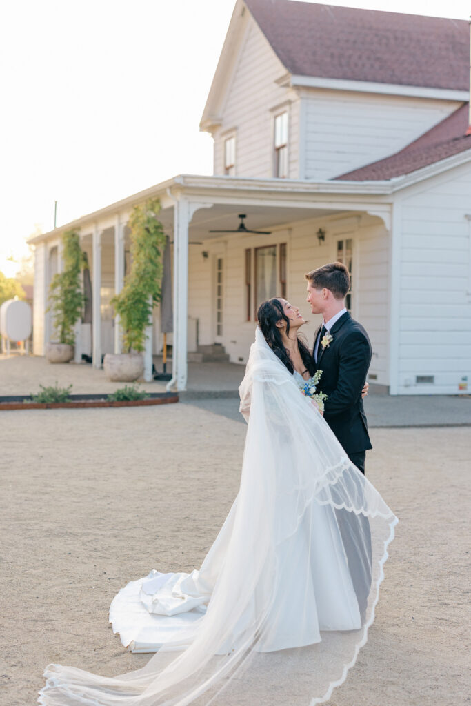A bride and groom share a kiss beside the white Victorian farmhouse at a Northern California wedding venue, the bride's cathedral veil trailing across the gravel drive, photographed by Suzanne Karp Photography