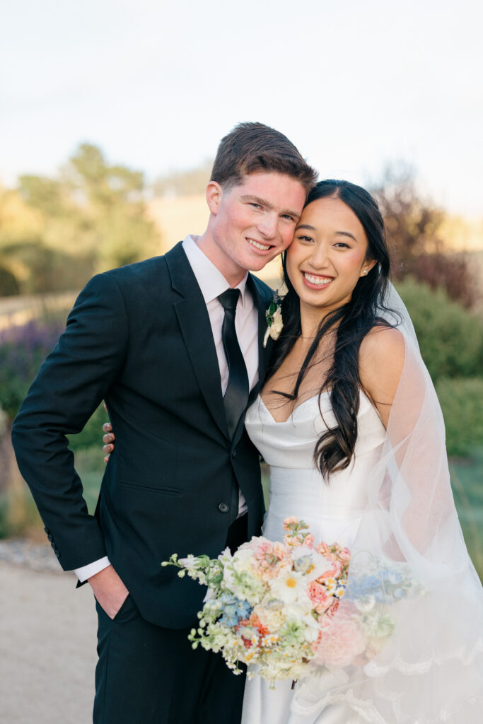 A bride and groom smile together during their golden hour portrait session in a Northern California vineyard, the bride holding a soft blush and white wildflower bouquet, photographed by Napa Valley wedding photographer Suzanne Karp