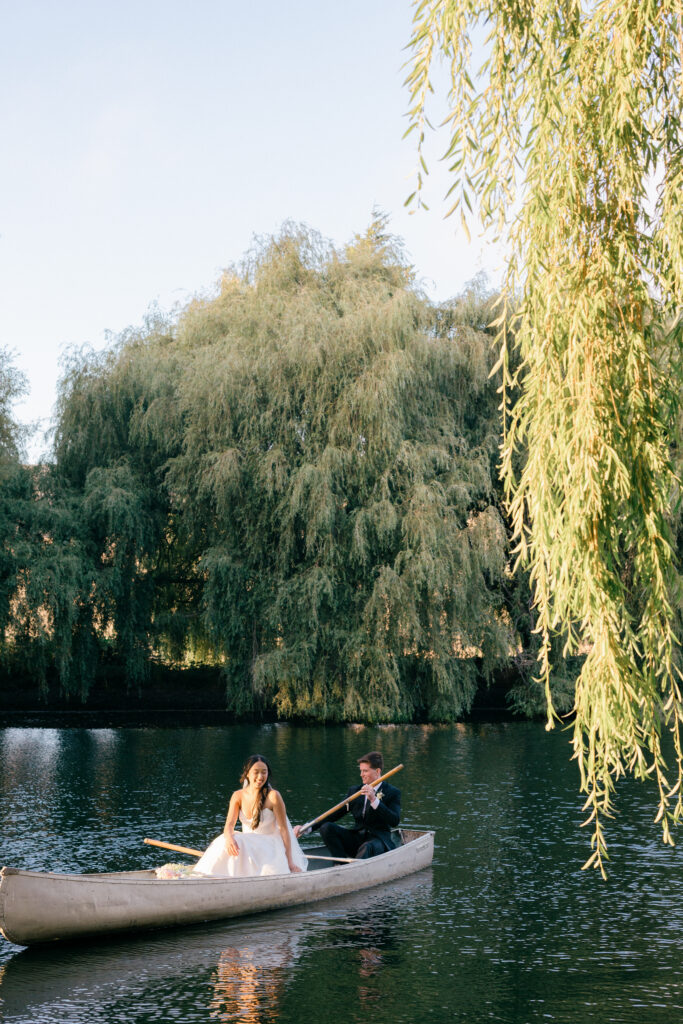 A bride and groom glide across a still lake in a wooden canoe beneath cascading weeping willow branches during their Northern California wedding portrait session, photographed by Napa Valley wedding photographer Suzanne Karp