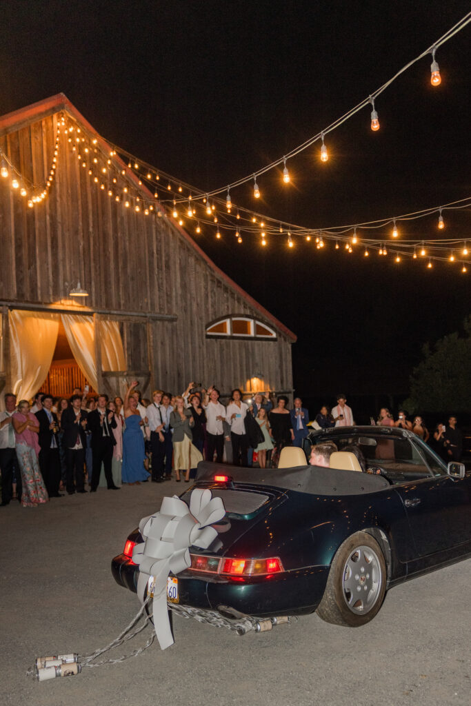 Wedding guests cheer and wave as a bride and groom make their grand exit in a navy vintage Porsche convertible decorated with a white bow beneath a canopy of string lights outside a red coastal barn wedding venue on the Sonoma Coast, photographed by Napa Valley wedding photographer Suzanne Karp