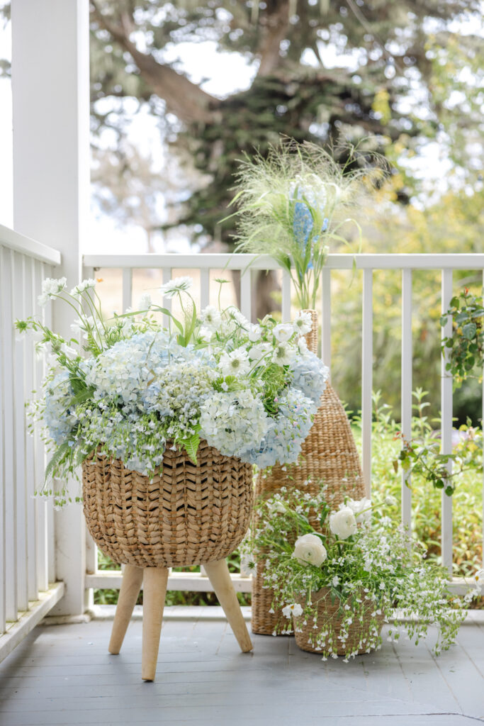 A woven basket arrangement of blue hydrangea, white blooms, and wispy grasses displayed on a wooden stand on the porch of a coastal Northern California wedding venue, photographed by Napa Valley wedding photographer Suzanne Karp