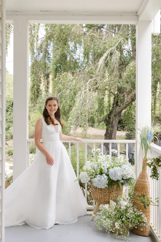 A bride in a simple strapless ballgown stands on the porch of a white Victorian farmhouse beside a woven basket arrangement of white hydrangea and wildflowers overlooking a coastal landscape, photographed by Napa Valley wedding photographer Suzanne Karp