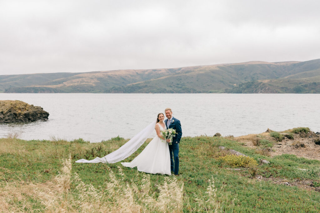 A bride and groom stand together in a wide landscape portrait on the rocky coastal shoreline with the calm bay and rolling hills stretching behind them and a red barn visible to the right during their Sonoma Coast wedding, photographed by Suzanne Karp Photography
