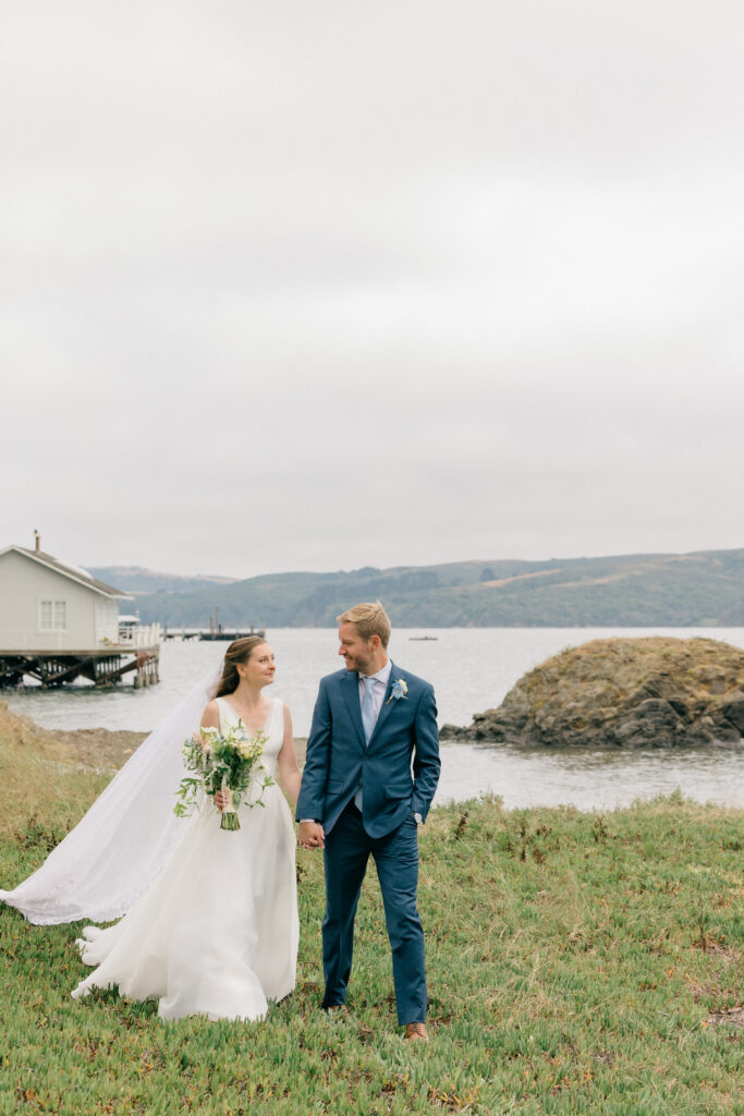 A bride and groom walk hand in hand along the grassy shoreline of a coastal bay with rolling hills and a wooden dock in the distance during their Northern California wedding portrait session, photographed by Suzanne Karp Photography