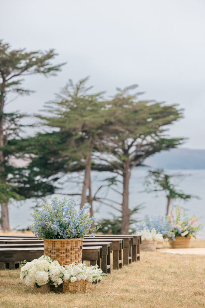 An outdoor coastal wedding ceremony setup with wooden pew benches, woven basket floral arrangements of white hydrangea and blue delphiniums, and a sweeping bay view beyond cypress trees on the Sonoma Coast, photographed by Napa Valley wedding photographer Suzanne Karp