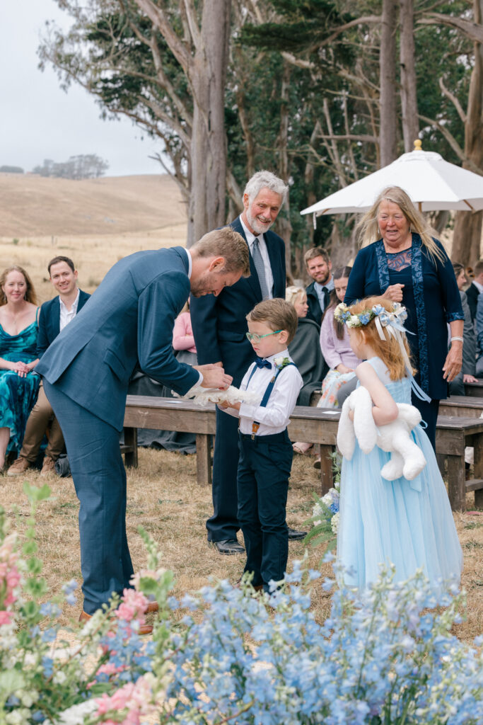 A groom in a navy suit kneels to greet a young ring bearer and flower girl in a blue tulle dress during an outdoor coastal wedding ceremony on the Sonoma Coast, with guests and rolling hills visible behind them, photographed by Suzanne Karp Photography