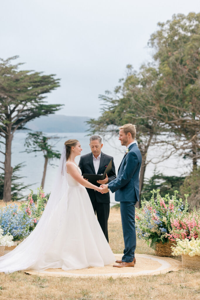 A bride and groom hold hands and exchange vows with their officiant during an outdoor coastal wedding ceremony surrounded by woven basket arrangements of colorful wildflowers and a bay view beyond the cypress trees on the Sonoma Coast, photographed by Napa Valley wedding photographer Suzanne Karp