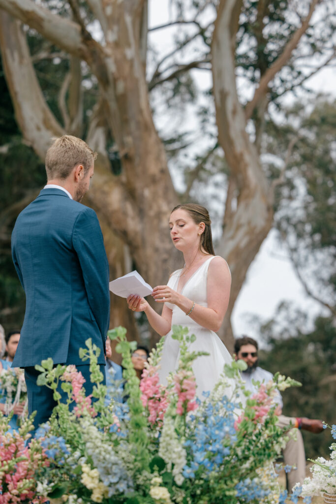 A bride reads her personal vows to her groom during an intimate outdoor coastal wedding ceremony on the Sonoma Coast, with lush pink and blue wildflower arrangements blooming in the foreground, photographed by Napa Valley wedding photographer Suzanne Karp