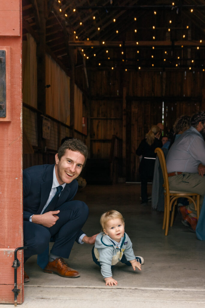 A candid moment of a groom in a navy suit crouching down to interact with a tiny toddler guest at the entrance of a warmly lit red barn wedding reception on the Sonoma Coast, photographed by Napa Valley wedding photographer Suzanne Karp