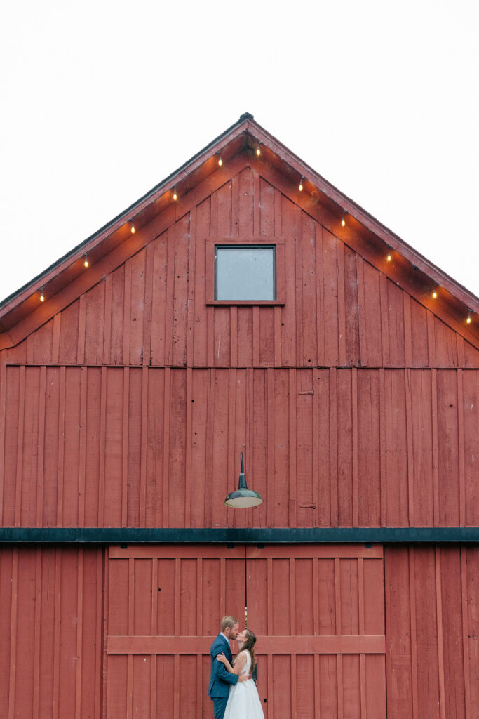 A bride and groom share a kiss against the dramatic red exterior of a coastal barn wedding venue, string lights lining the roofline above them, photographed by Suzanne Karp Photography