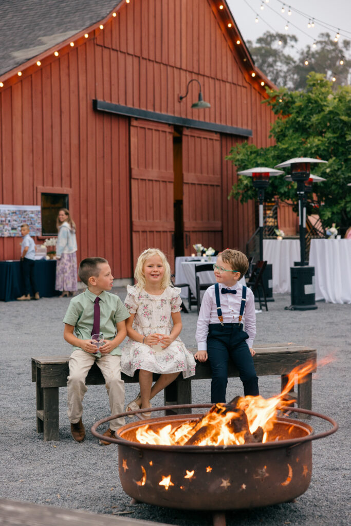 Three children in formal wedding attire sit together beside a glowing fire pit during an outdoor coastal barn wedding reception on the Sonoma Coast, photographed by Napa Valley wedding photographer Suzanne Karp