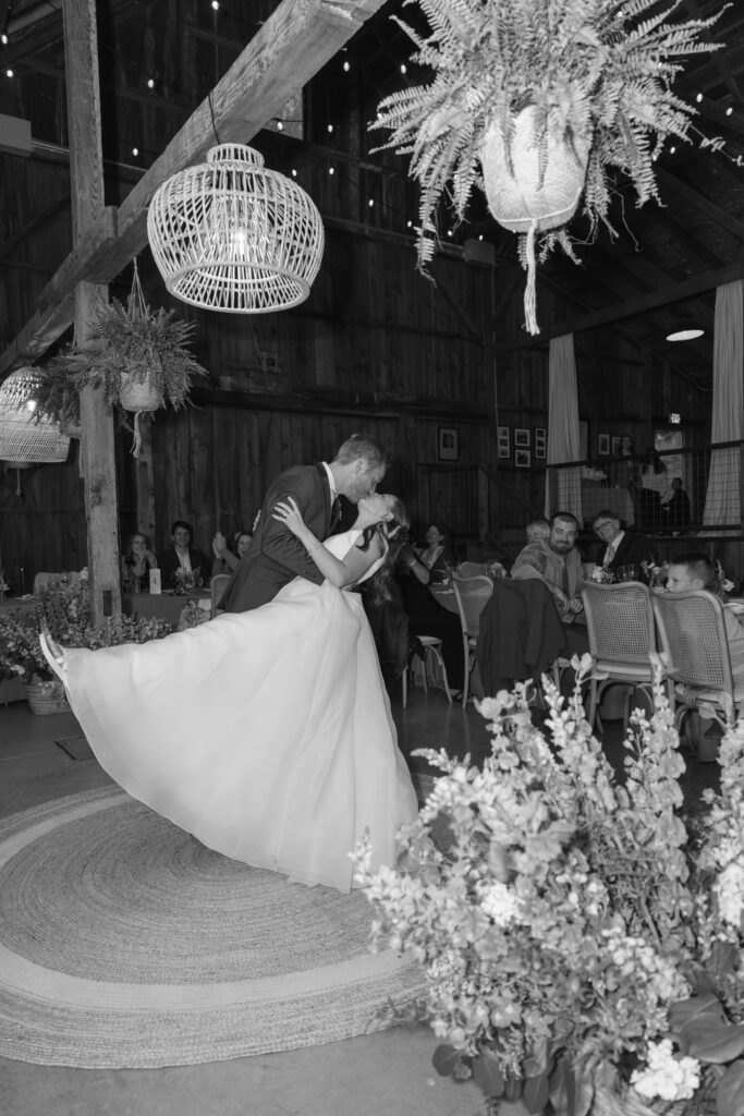 A black and white photograph of a groom dipping his bride during their first dance inside a rustic barn wedding venue, a woven pendant chandelier and hanging ferns above them and guests watching from candlelit tables, photographed by Suzanne Karp Photography