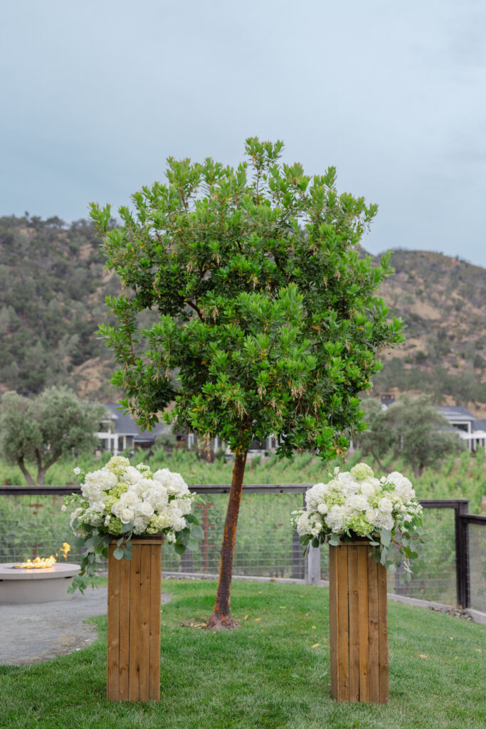 Elopement ceremony setup at Four Seasons Napa Valley, two white floral arrangements on wooden pedestals beside a fire pit with vineyard and hills beyond