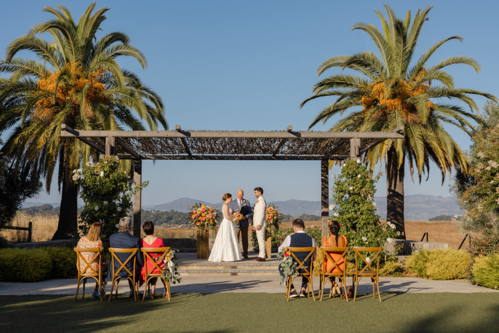 Couple exchanges vows beneath the pergola at Carneros Resort and Spa, framed by date palms and coral florals, with a handful of guests seated on the lawn