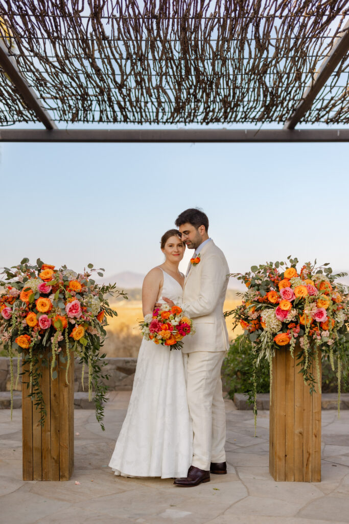 Couple embraces beneath the woven pergola at Carneros Resort and Spa, framed by coral and orange floral arrangements, during their Napa Valley elopement