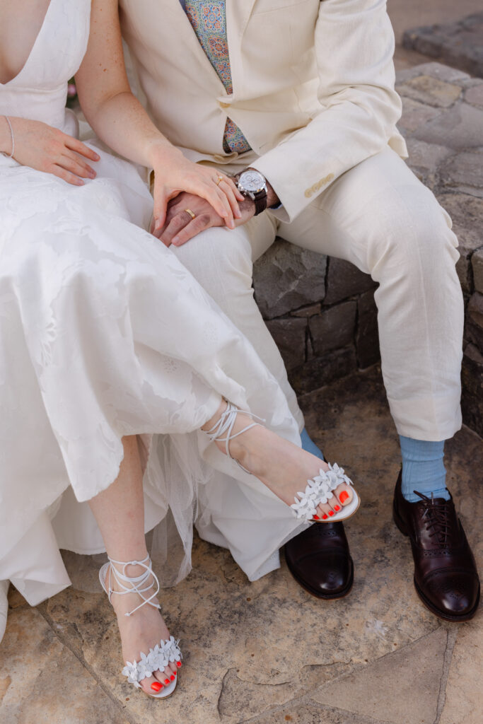 Bride and groom sit side by side holding hands, her lace-up heels beside his brogues, during their Napa Valley elopement
