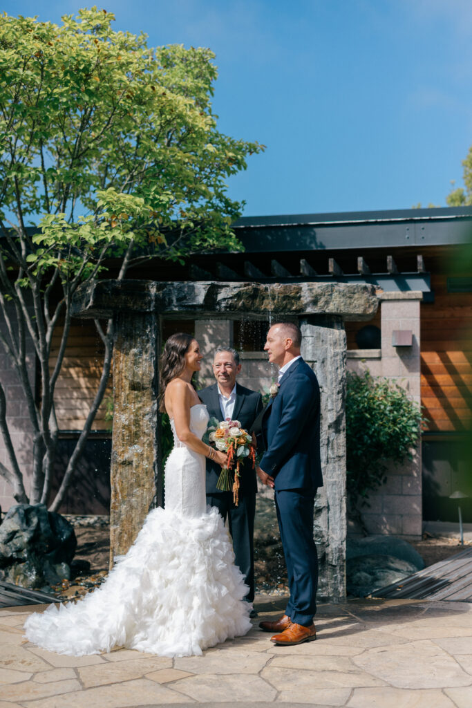 Bride in a ruffled gown and groom in navy face each other as their officiant begins the ceremony beneath a stone and timber arbor