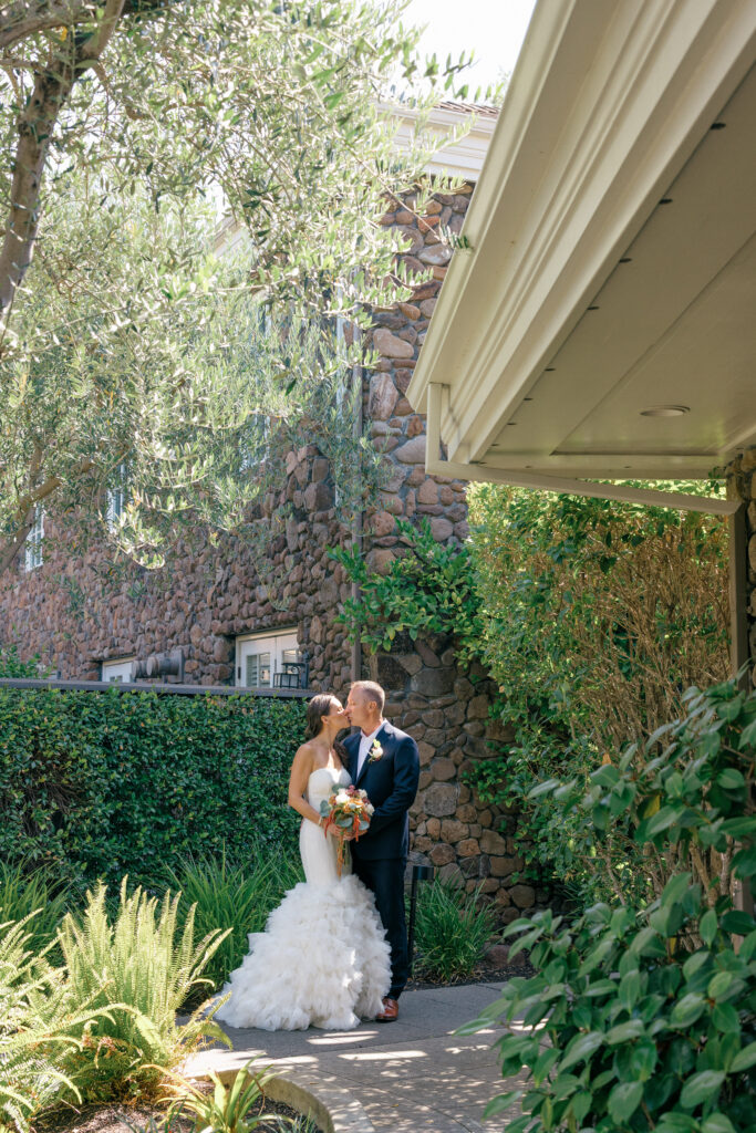 Couple shares a kiss on a garden path beside a stone wall and olive tree before their Napa Valley elopement ceremony