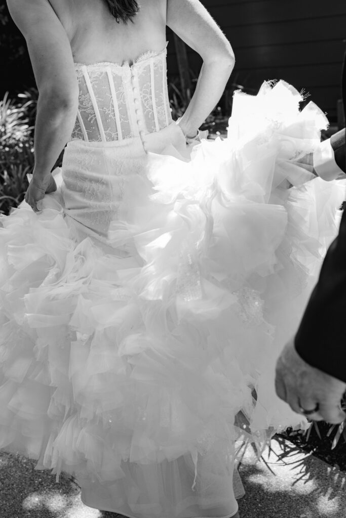 Bride lifts the tiered tulle skirt of her gown, her groom's hand and wedding band at her side, in black and white