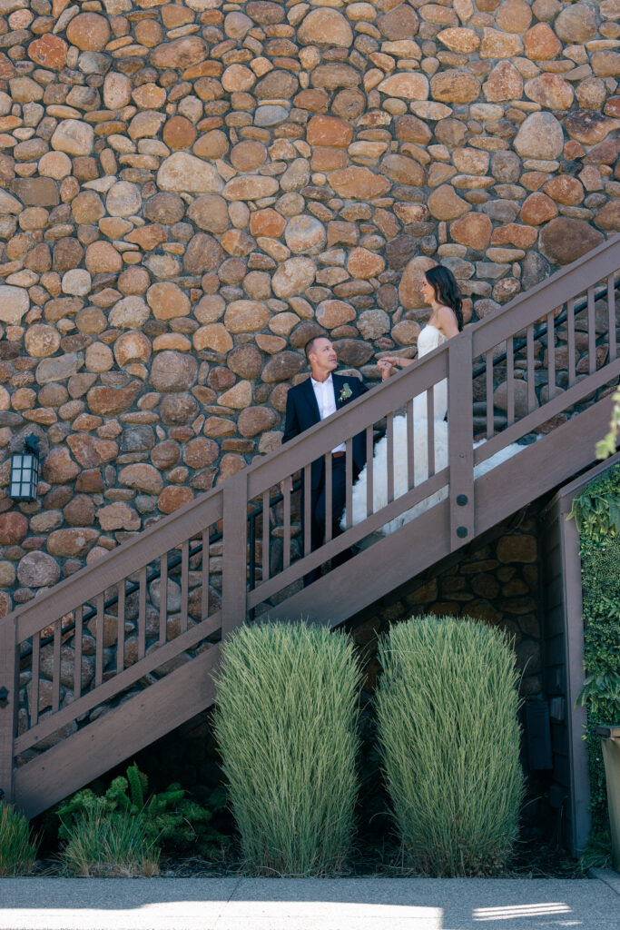 Groom leads his bride down a wooden staircase against the river rock wall at Bardessono, framed by ornamental grasses